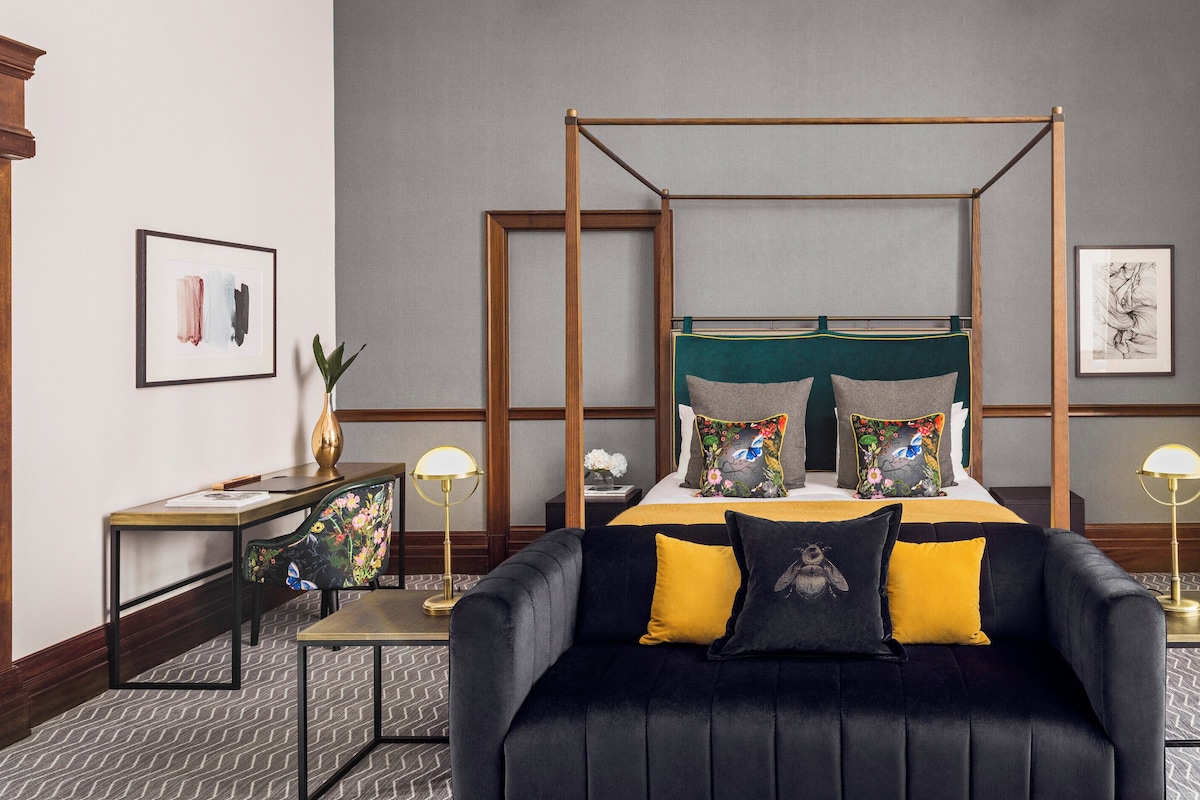 Habitació d'hotel amb un llit amb cortines, un sofà negre amb coixins grocs, una taula petita i un escriptori amb una làmpada.