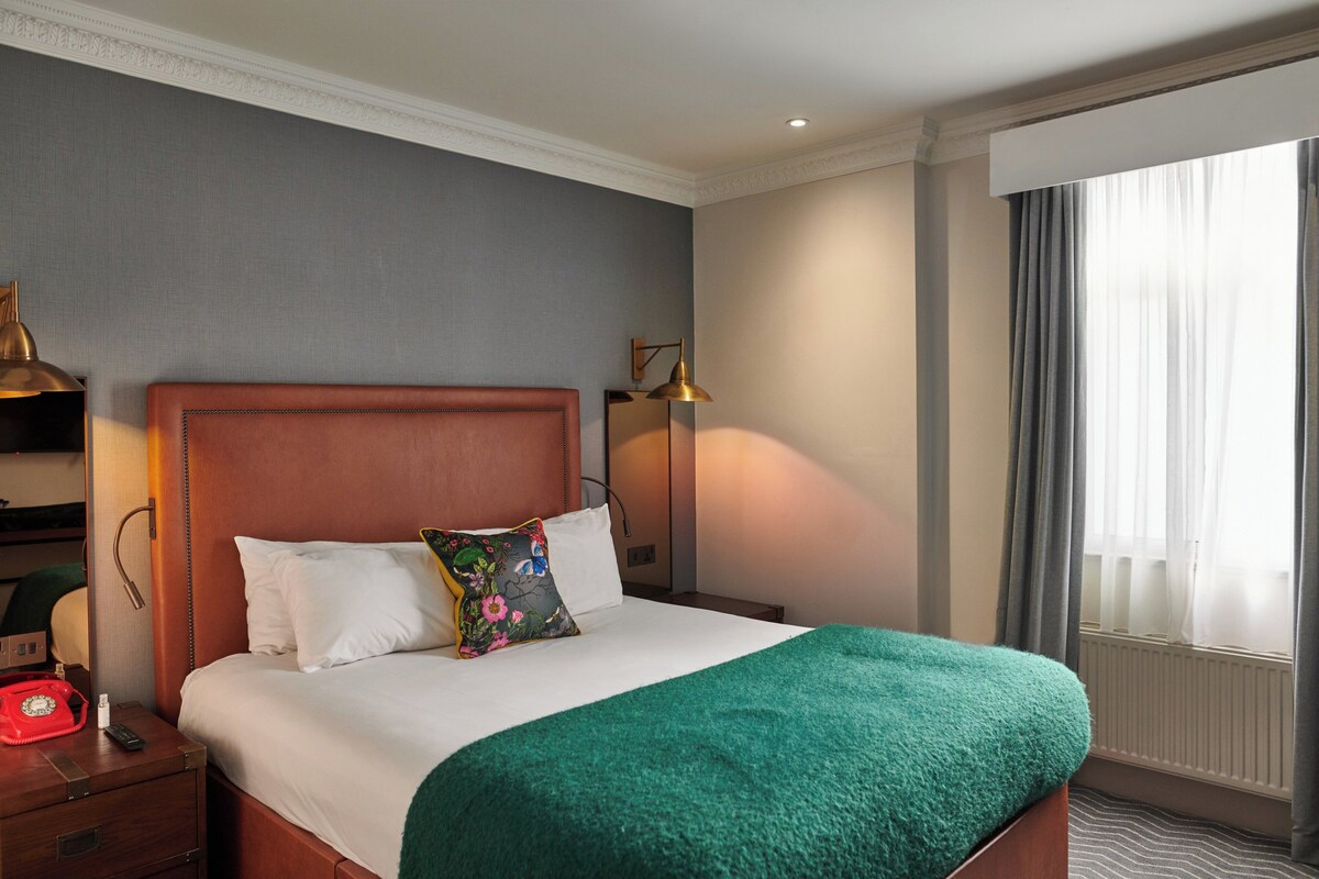 A hotel room with a large bed, a nightstand with a red telephone, a lamp, and a window with curtains.