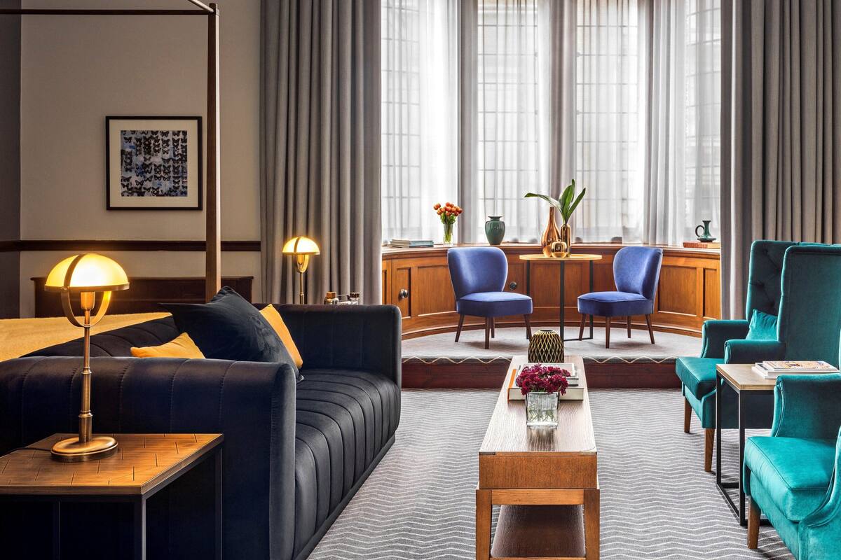 A modern hotel room with a dark blue sofa, teal armchairs, a wooden coffee table, and a desk with chairs.