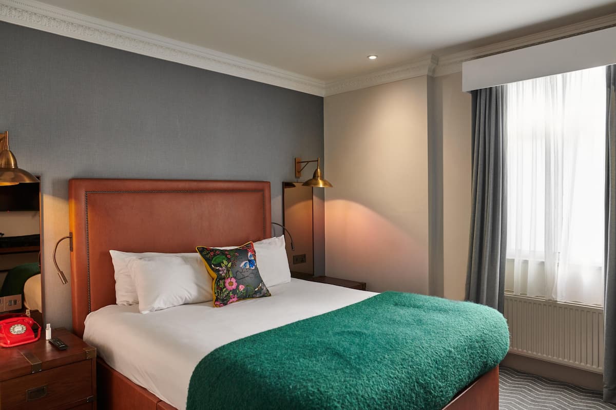 Une chambre d’hôtel avec un lit, une table de chevet sur laquelle se trouve un téléphone rouge, une lampe et une fenêtre avec des rideaux.