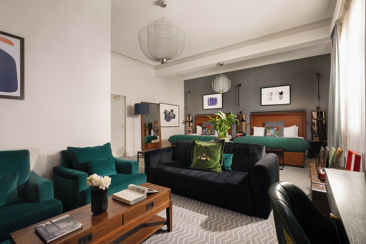 A modern hotel room with a green sofa, a wooden coffee table, a bed with a green headboard, and a large window with curtains.