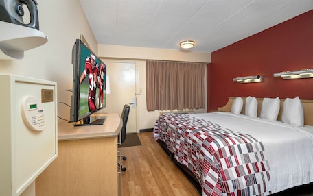 A hotel room with a bed, a desk with a television, a desk lamp, a wall-mounted light, a door with a curtain, and a wall-mounted shelf.