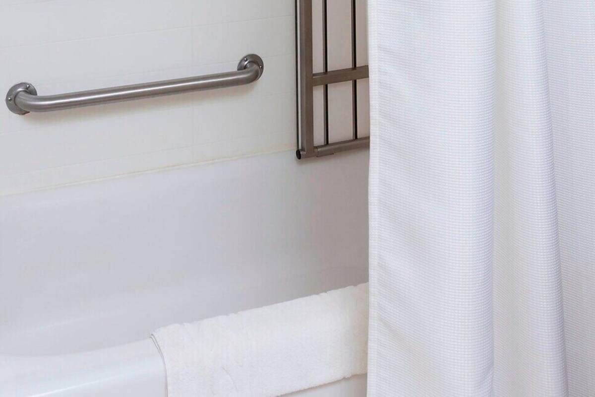 Une salle de bain avec une baignoire blanche, une barre d’appui en métal et un rideau de douche blanc.