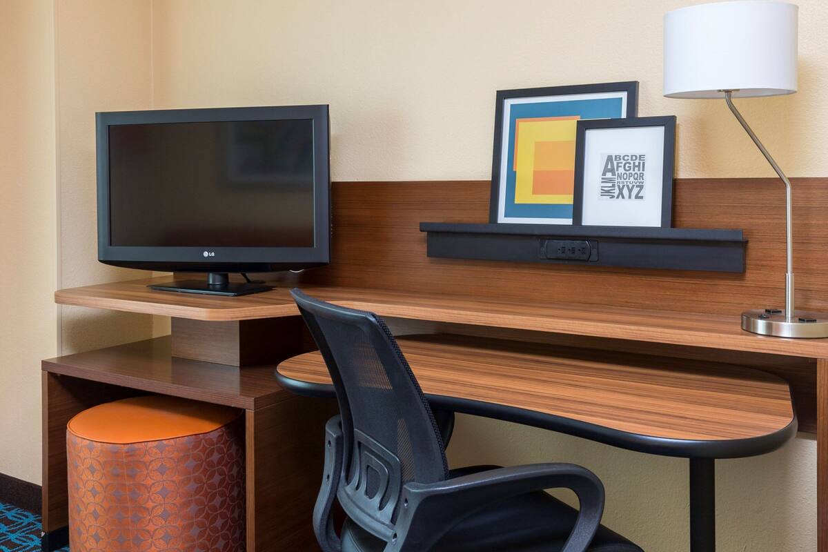 Ein Hotelarbeitsplatz mit Schreibtisch, Stuhl, Computermonitor, Lampe und gerahmtem Kunstwerk.