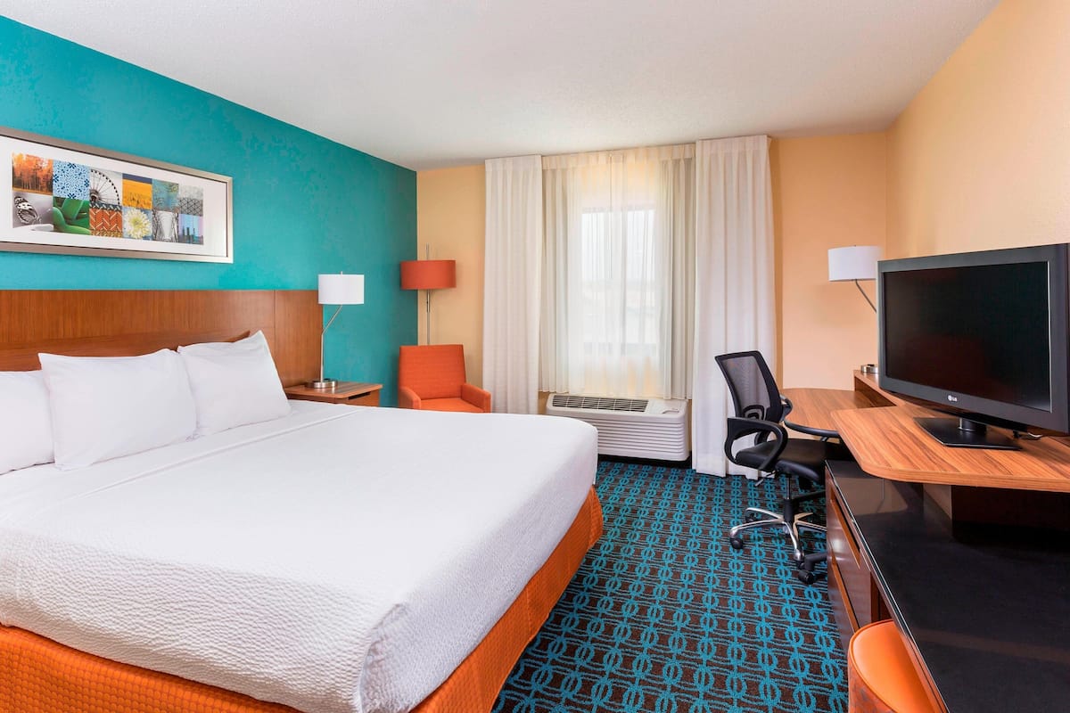 A hotel room with a large bed, a desk with a TV, a chair, an air conditioner, and a painting on the wall.