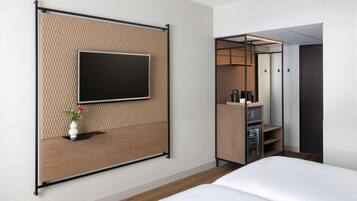 Ein modernes Hotelzimmer mit einem Flachbildfernseher an einer strukturierten Wand, einem Nachttisch aus Holz mit Blumen in einer Vase, einem eingebauten Minikühlschrank und einem Kleiderschrank mit Spiegel.