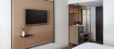 Ett modernt hotellrum med en platt-TV som är monterad på en vägg med en intressant yta, ett träbord vid sängen med en vas med blommor, ett inbyggt minikylskåp och en garderob med en spegel.