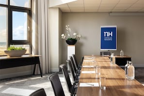 A conference room with a long wooden table, chairs, a potted plant, a banner, and a view of the ocean.