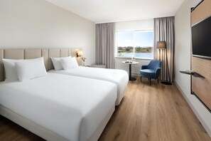 Habitación de hotel con dos camas, un escritorio, una silla, un televisor y una ventana con vista a un lago.