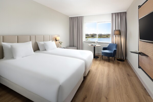 A hotel room with two beds, a desk, a chair, a TV, and a window with a view of a lake.