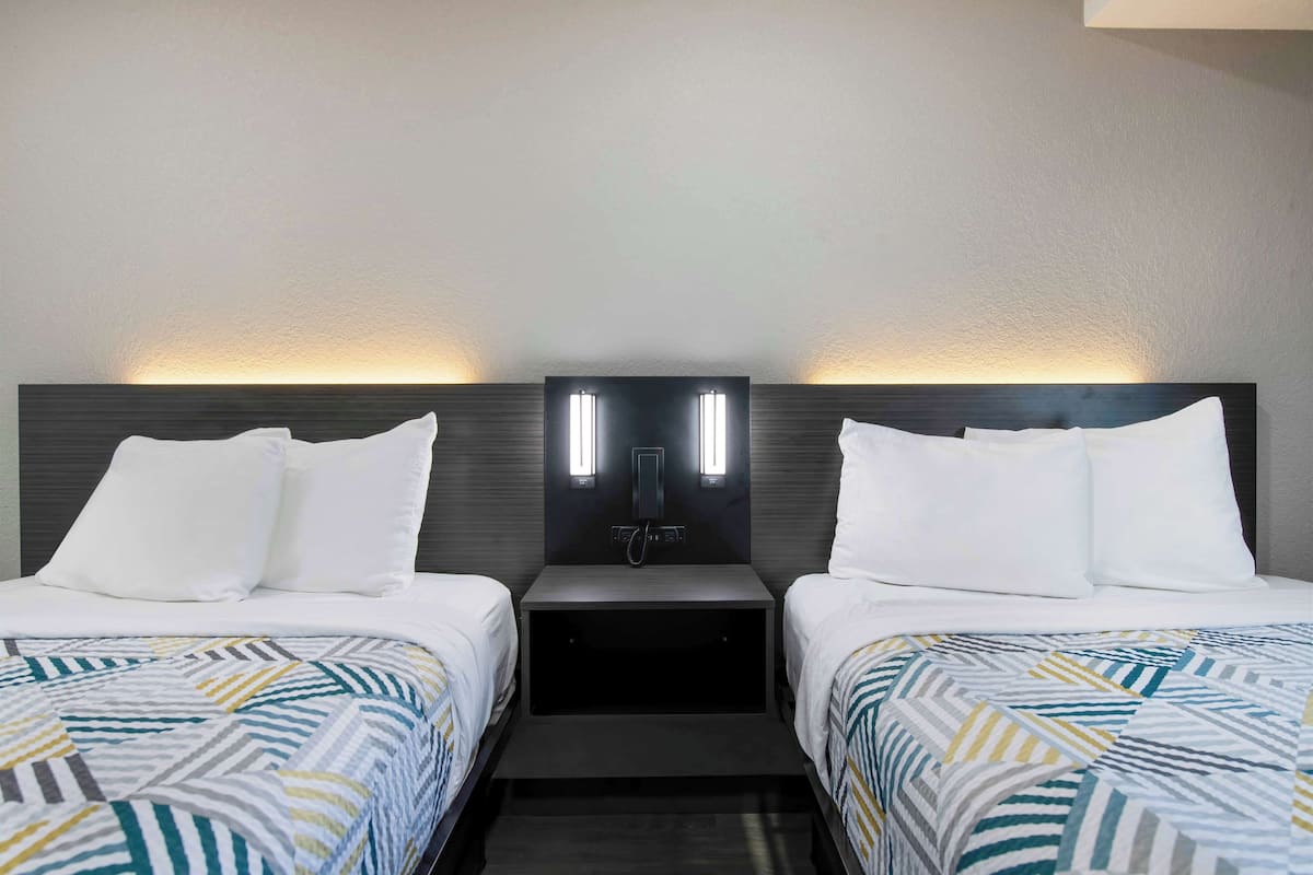 Habitación de hotel con dos camas, cada una con ropa de cama blanca y una colcha estampada. Entre las camas hay una mesita de noche con una lámpara moderna.