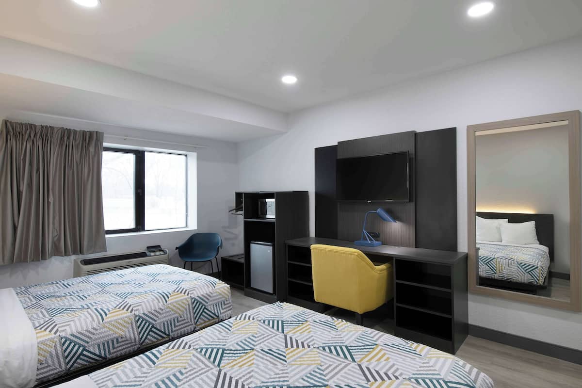 Habitación de hotel con dos camas, un escritorio, una silla, un televisor, un espejo y una ventana con cortinas.