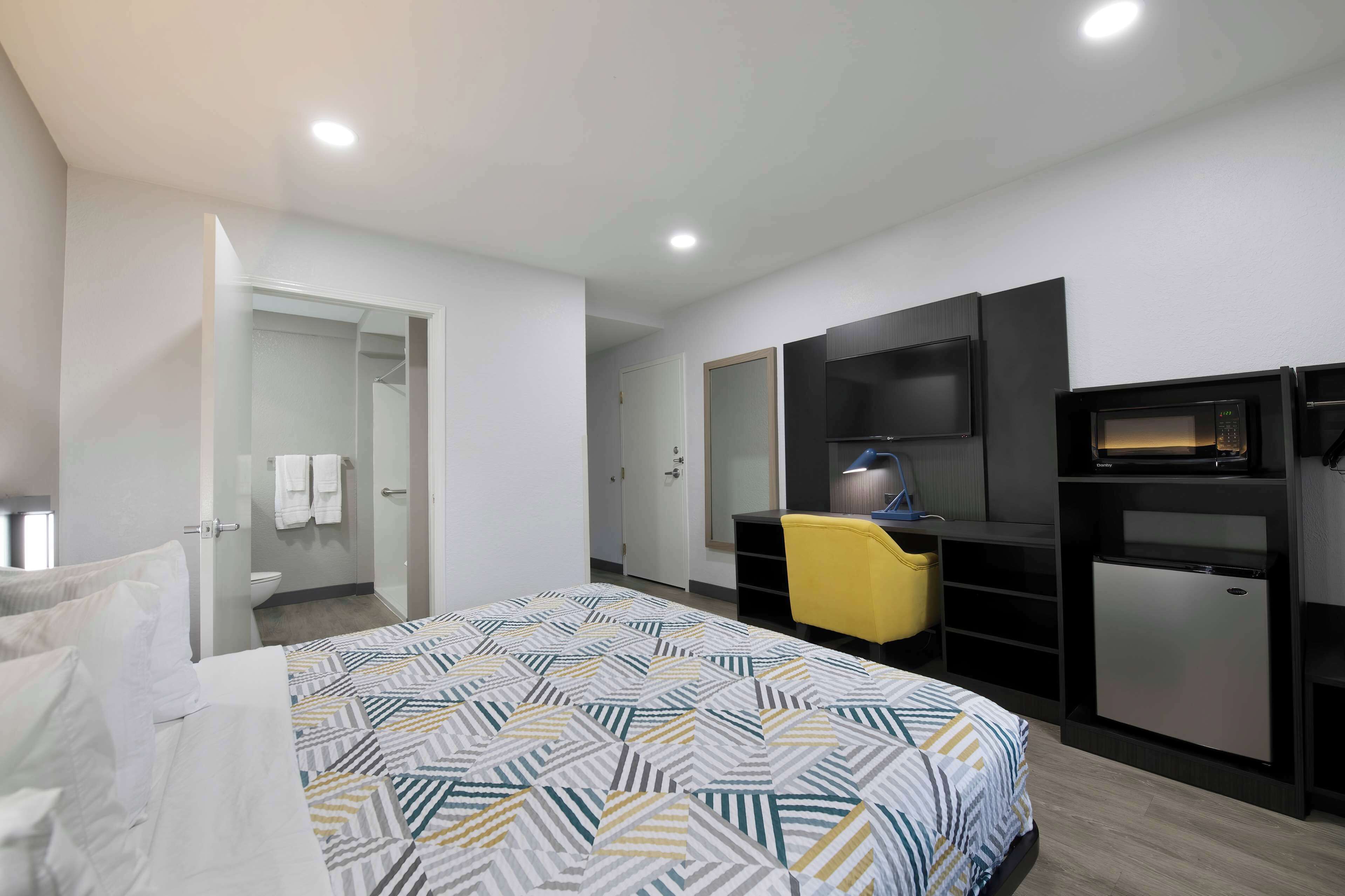Una habitación de hotel moderna con una cama, un escritorio, una silla, un televisor y un refrigerador pequeño.