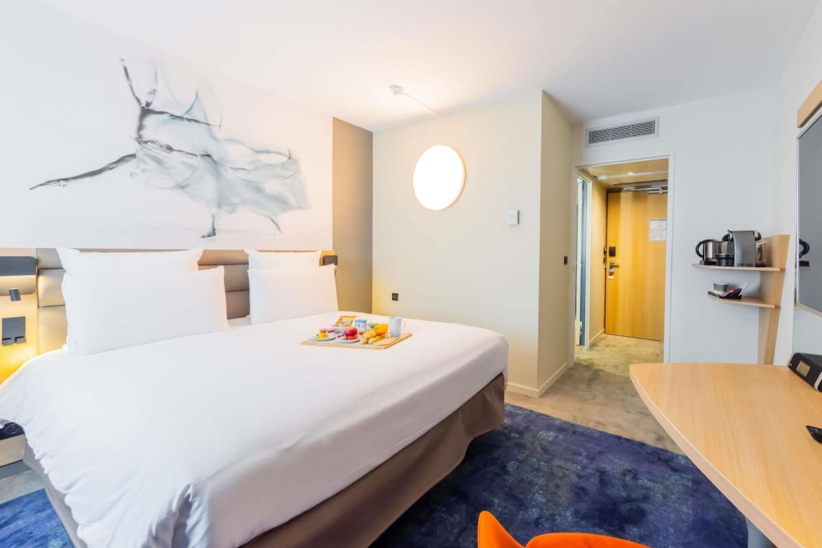 A hotel room with a bed, a round wall light, a small table, and a door leading to a bathroom.