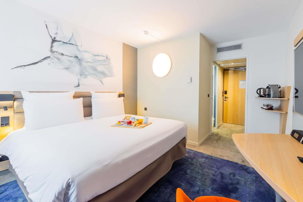 Une chambre dâhĂŽtel avec un lit, un applique ronde au mur, une petite table avec une bouilloire et une porte donnant sur une salle de bain.
