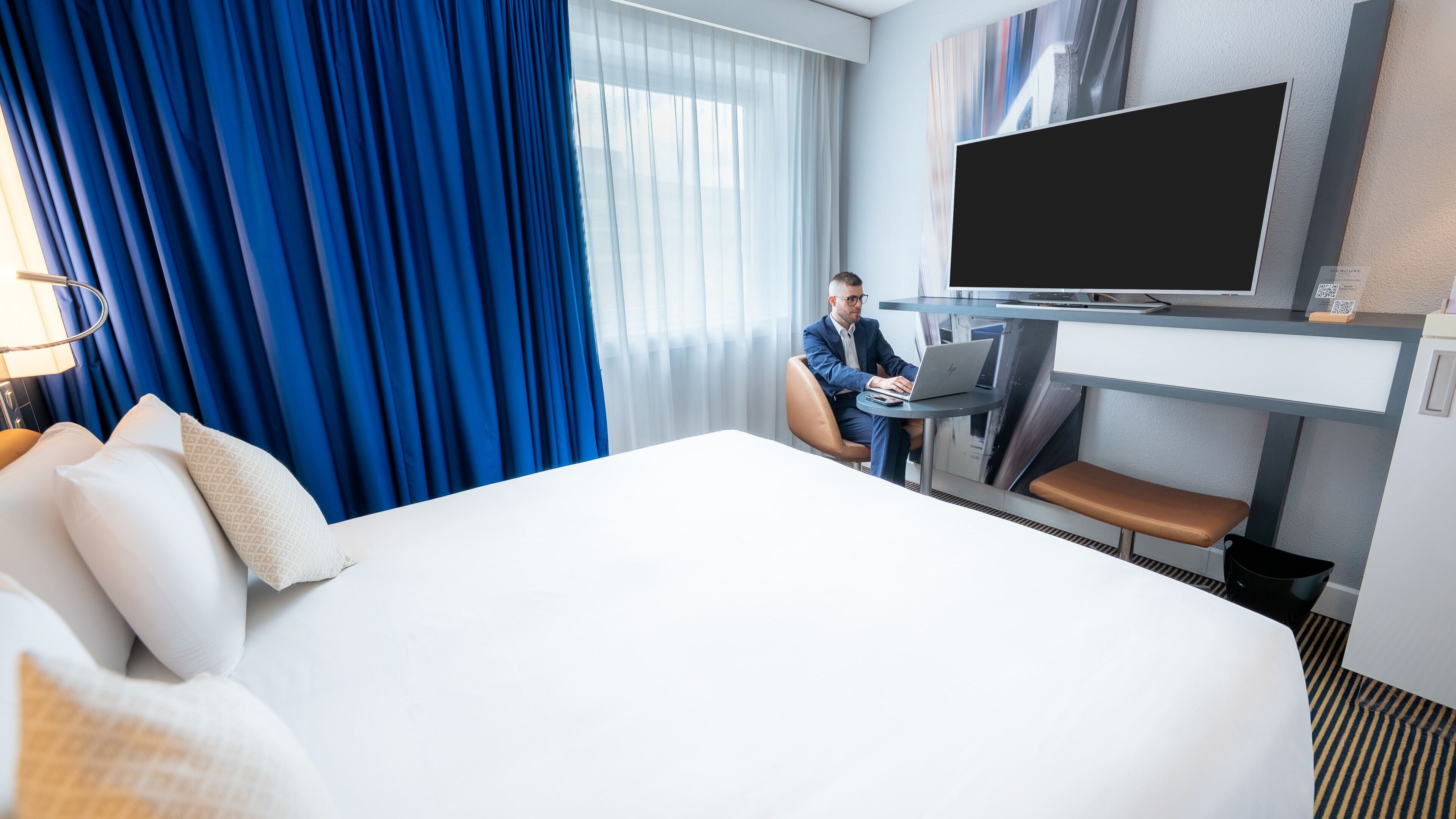 A hotel room with a large bed, a desk with a laptop, a flat-screen TV, and a large window with blue curtains.