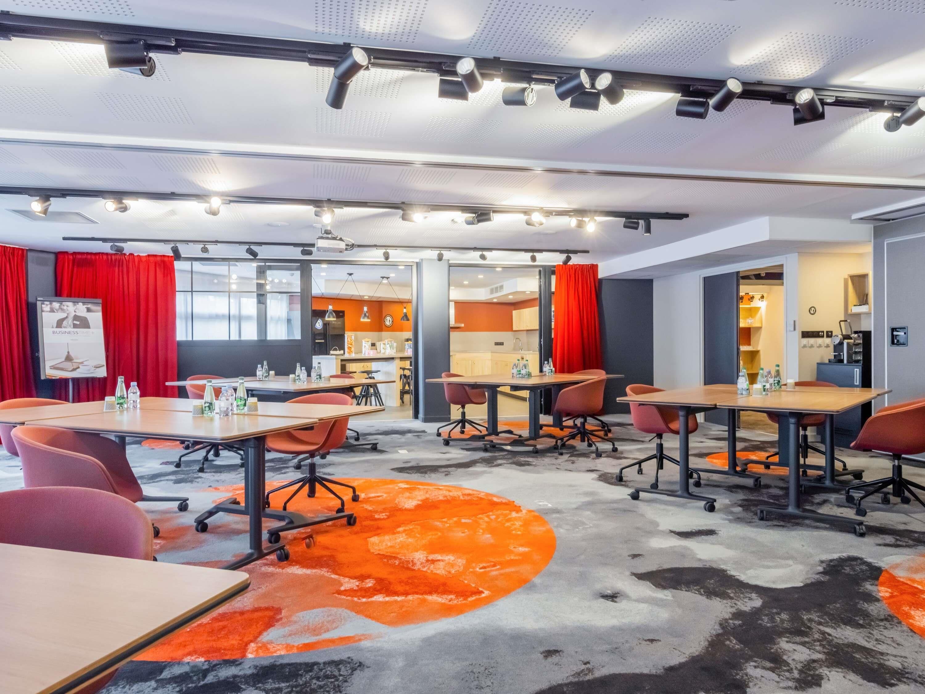 A modern conference room with tables, chairs, and a kitchen area visible through an open door.