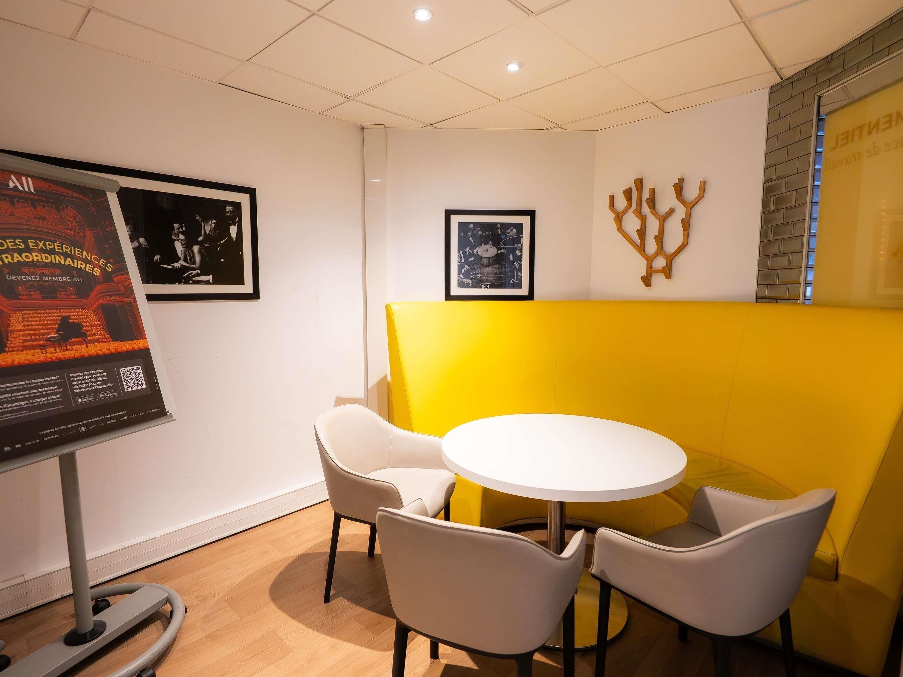 A modern meeting room with a white table, four chairs, a standing desk, and a poster on the wall.