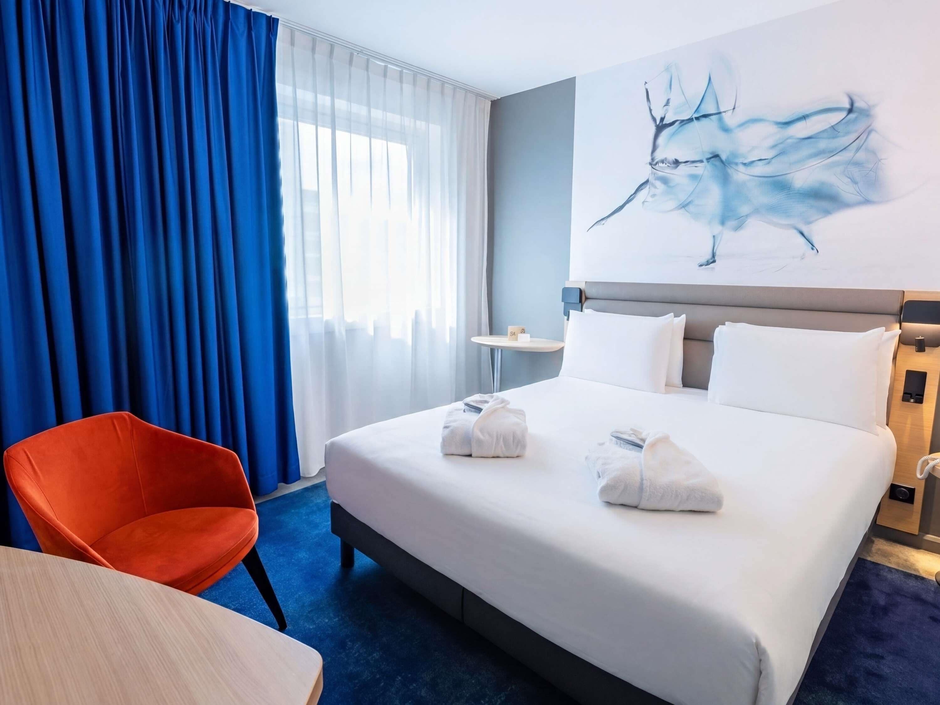 Hotellihuone, jossa on sänky, tuoli, pöytä, seinällä taulu ja ikkuna, jossa on verhot.