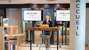 Un área de recepción con un escritorio de madera, una computadora portátil y una persona sentada. Hay dos letreros grandes que dicen 'MERCURE HOTELS' y un pilar con la palabra 'ACCUEL'.