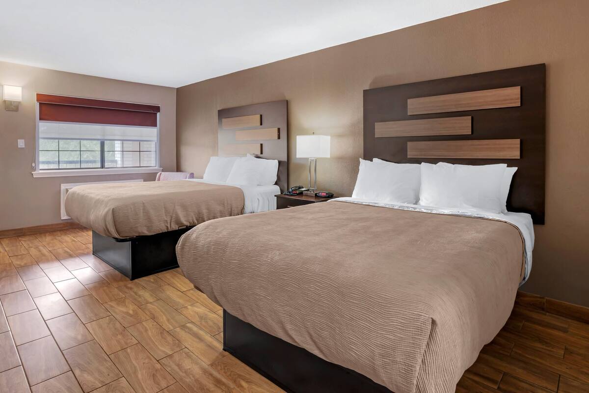 A hotel room with two beds, a bedside table with a lamp, a window with blinds, and a wooden floor.