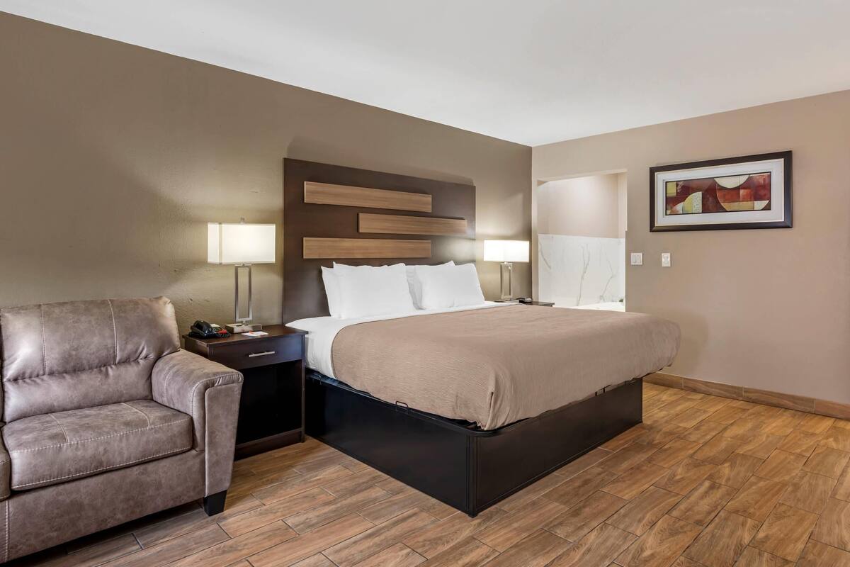 A modern hotel room with a bed, a chair, a nightstand, and a wall-mounted shelf.