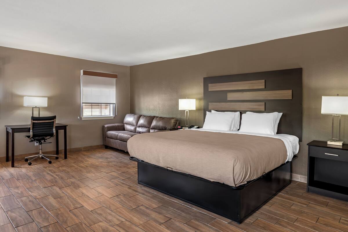 A modern hotel room with a large bed, a leather sofa, a desk with a chair, and two bedside tables with lamps.