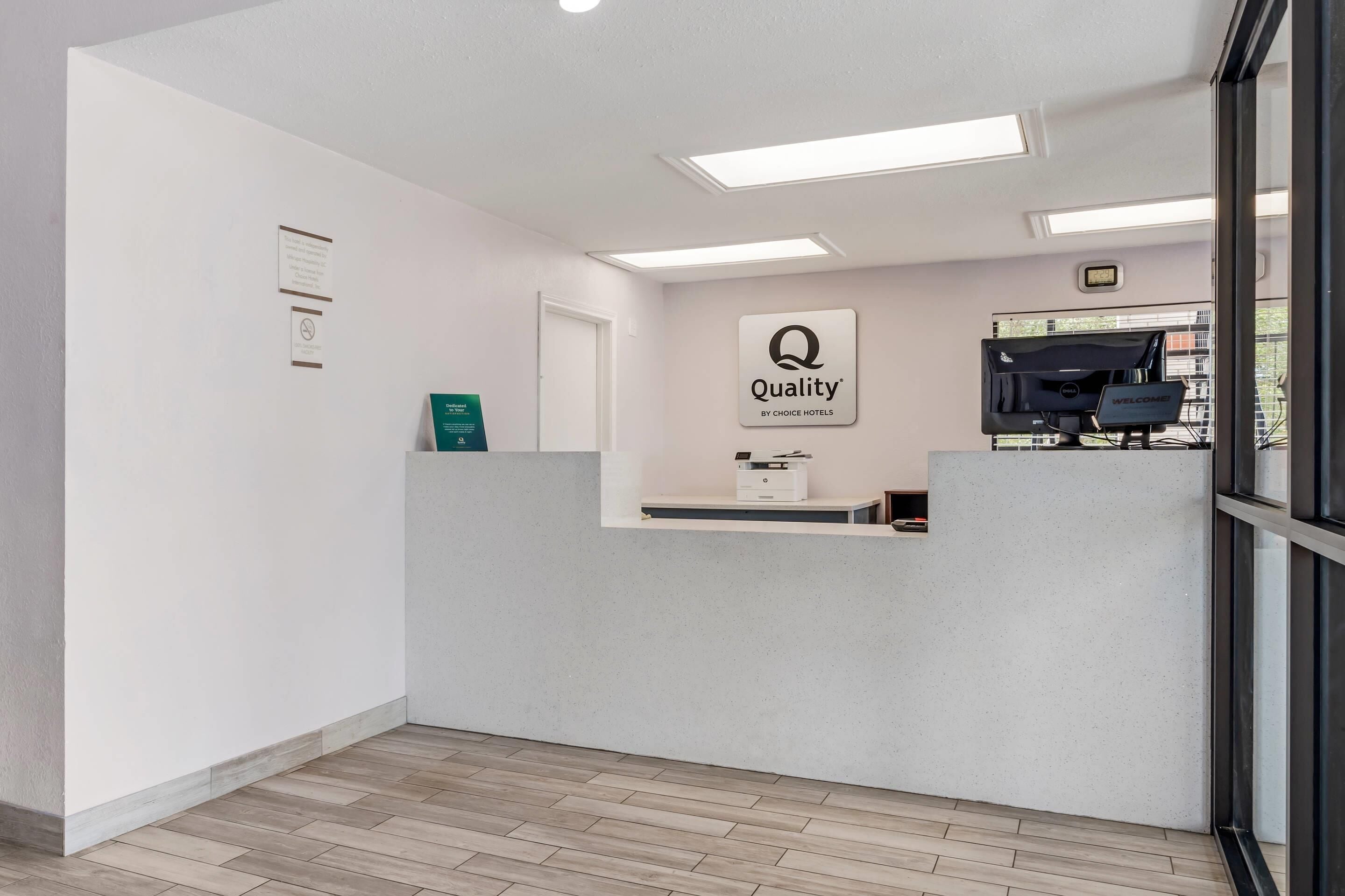A reception area with a 'Quality by Choice Hotels' sign, a reception desk, a digital clock, and a window with blinds.