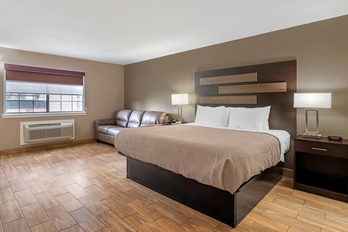A hotel room with a bed, a sofa, two bedside lamps, a heater, and a window with blinds.