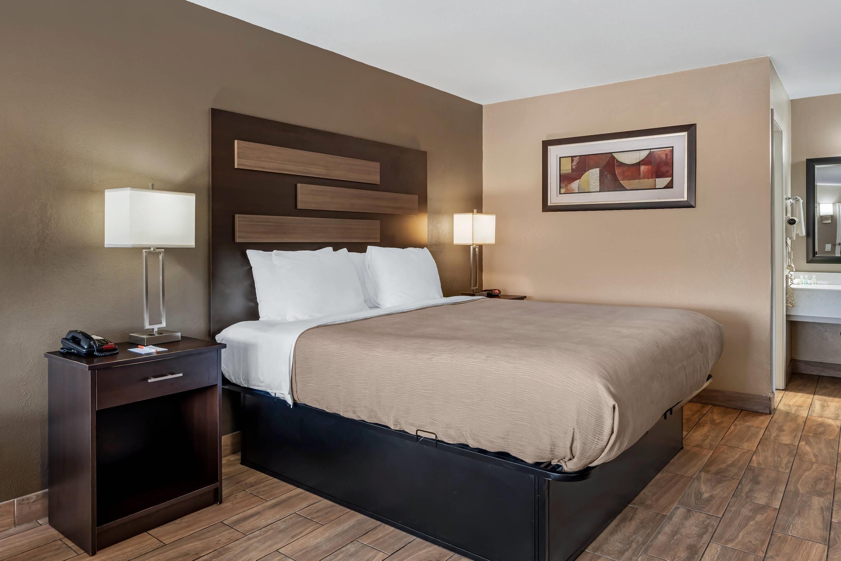 A hotel room with a bed, bedside tables, lamps, a framed picture, and a bathroom visible through an open door.