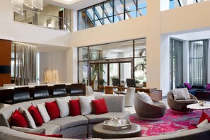 Eine großzügige Lobby mit einer gebogenen grauen Couch, verschiedenen Sitzgelegenheiten, einem Empfangstresen und großen Fenstern mit Blick ins Freie.