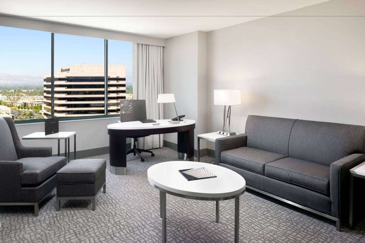 Una habitación de hotel moderna con un sofá gris, una pequeña mesa redonda, un escritorio con una silla y un amplio ventanal con vistas a la ciudad.