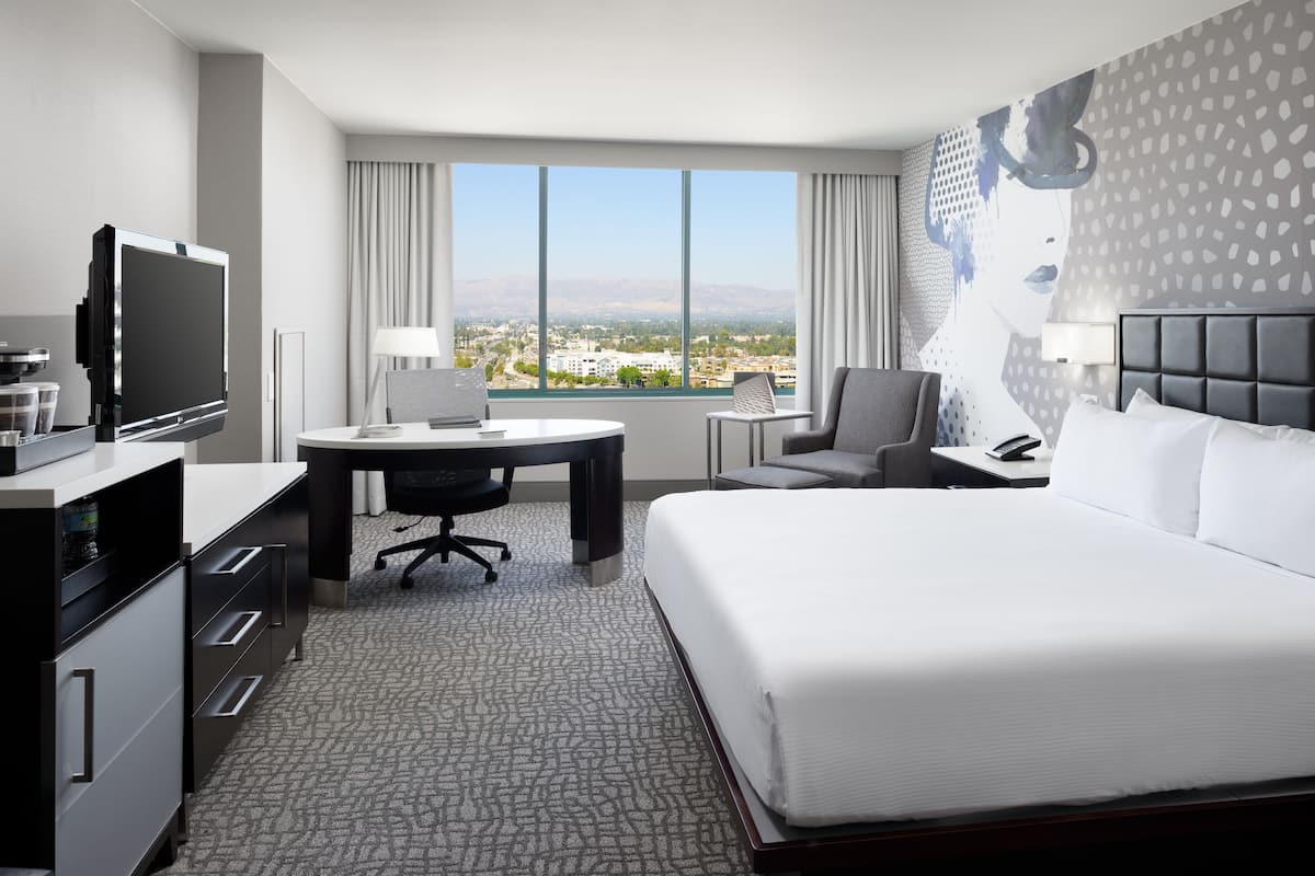 Habitación de hotel con una cama grande, un escritorio con ordenador, televisión y vistas a un paisaje urbano a través de amplios ventanales.