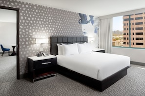 Une chambre d’hôtel moderne avec un grand lit, une table de chevet avec une lampe, un bureau avec une chaise et une vue sur la ville par la fenêtre.