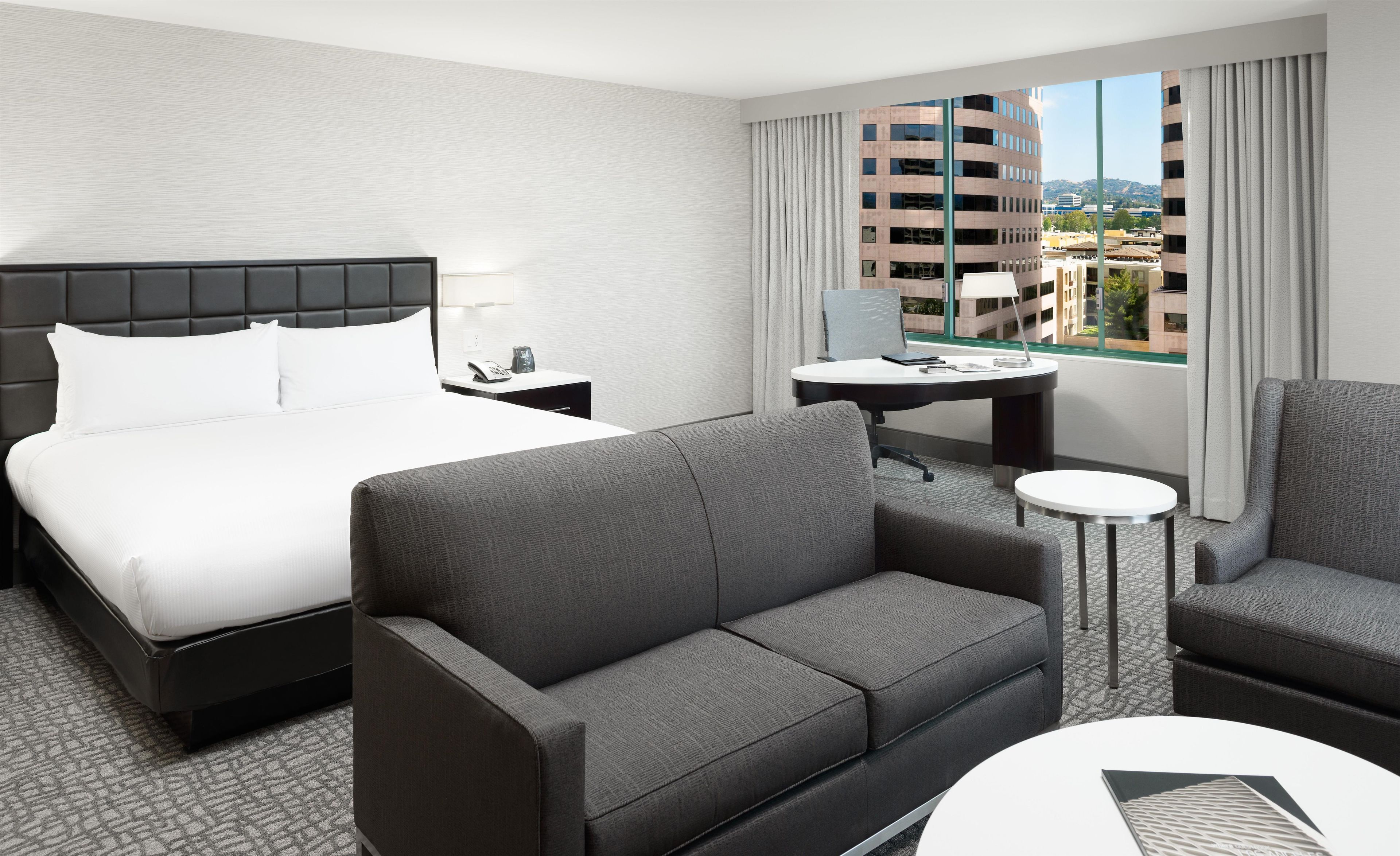 A hotel room with a bed, a sofa, a desk, and a view of buildings.