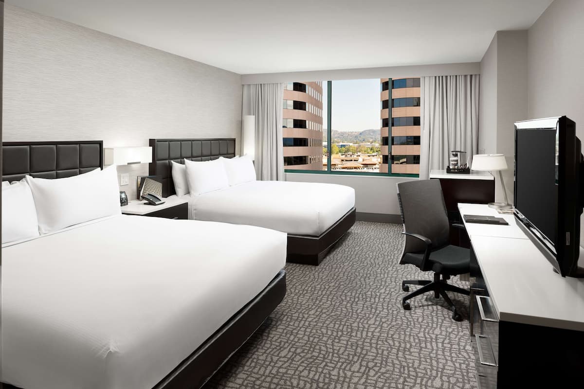 Habitación de hotel con dos camas, un escritorio con ordenador, televisor y ventana con vistas a la ciudad.