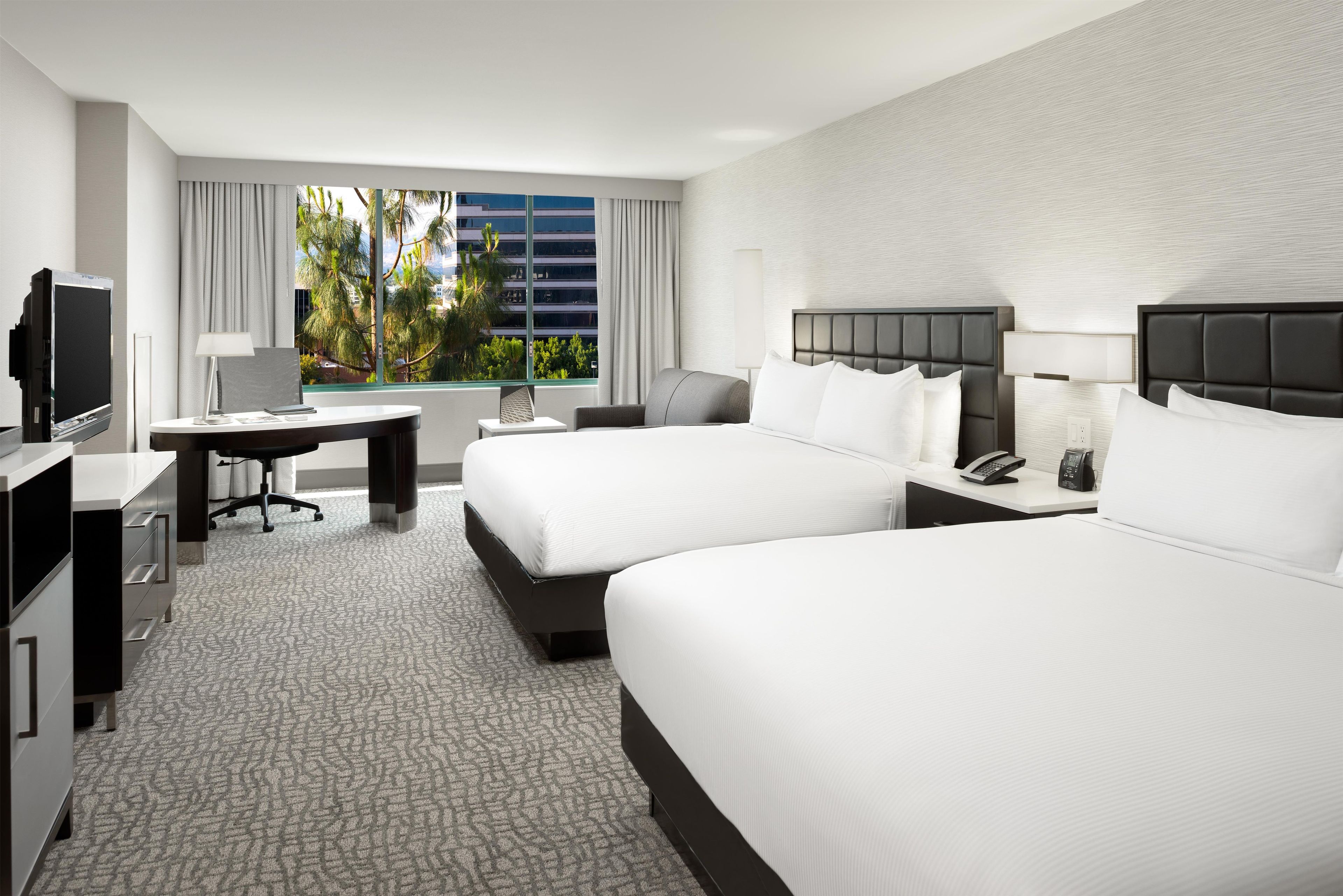 A hotel room with two beds, a desk, a TV, and a window with a view of palm trees.