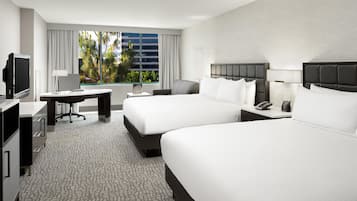 A hotel room with two beds, a desk, a TV, and a window with a view of palm trees.