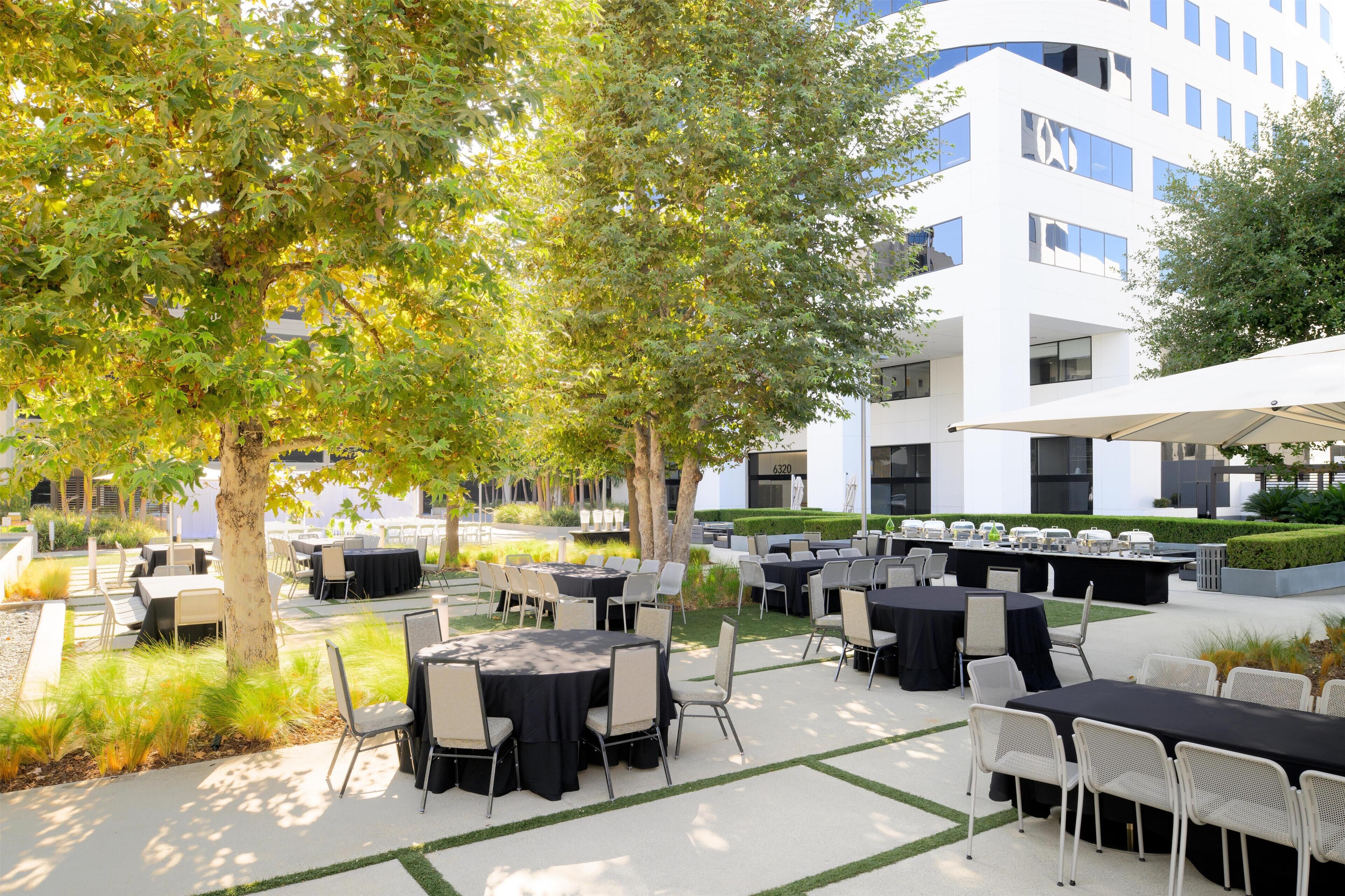 Un patio al aire libre con mesas y sillas, rodeado de árboles y un edificio moderno al fondo.