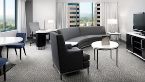 Una habitación de hotel moderna con un sofá curvo, un televisor de pantalla plana, una mesa de comedor y vistas a un paisaje urbano a través de amplios ventanales.