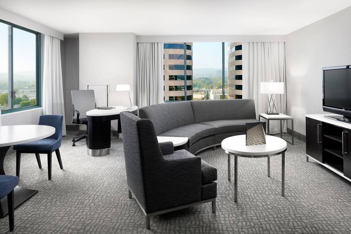 Una habitación de hotel moderna con un sofá curvo, un televisor de pantalla plana, una mesa de comedor y vistas a un paisaje urbano a través de amplios ventanales.