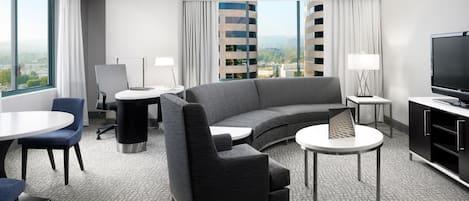 A modern hotel room with a curved sofa, a flat-screen TV, a dining table, and a view of a cityscape through large windows.