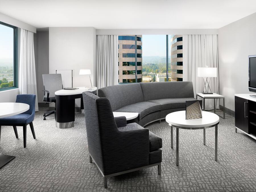 A modern hotel room with a curved sofa, a flat-screen TV, a dining table, and a view of a cityscape through large windows.
