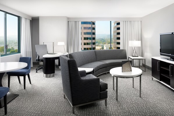 Quarto de hotel moderno com sofá curvo, TV de tela plana, mesa de jantar e vista para a cidade através de amplas janelas.