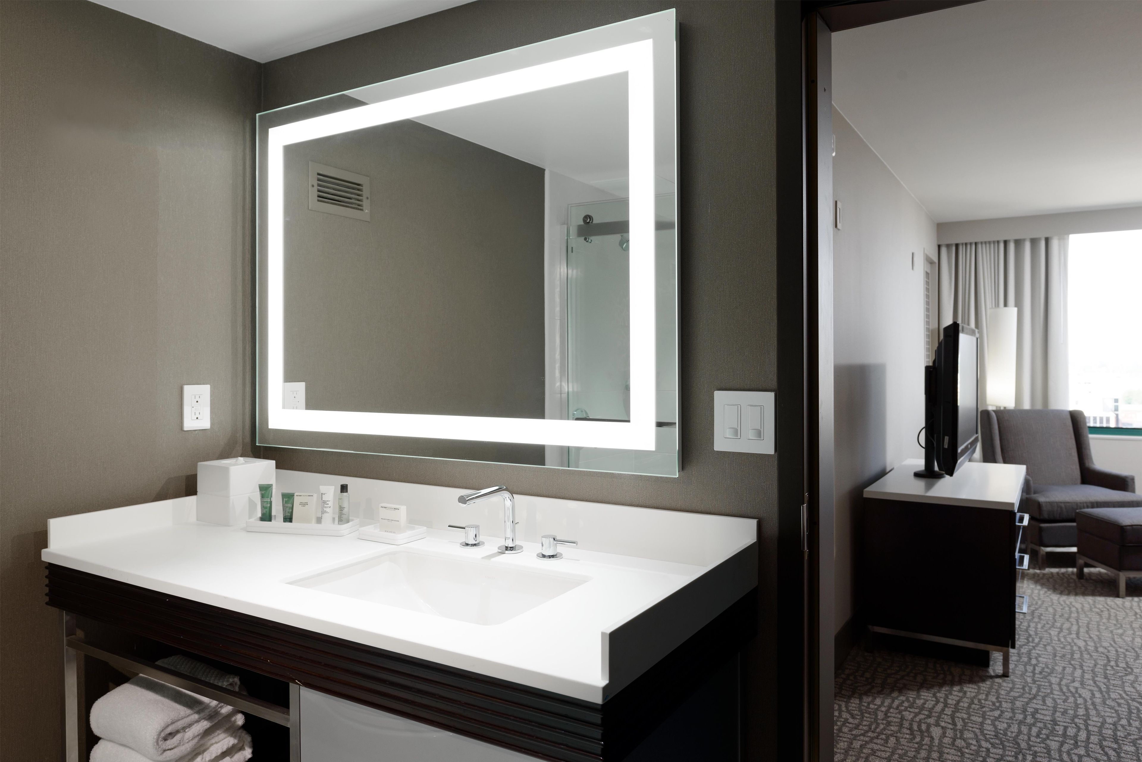 A hotel bathroom with a large mirror, a white sink, and a dark vanity.