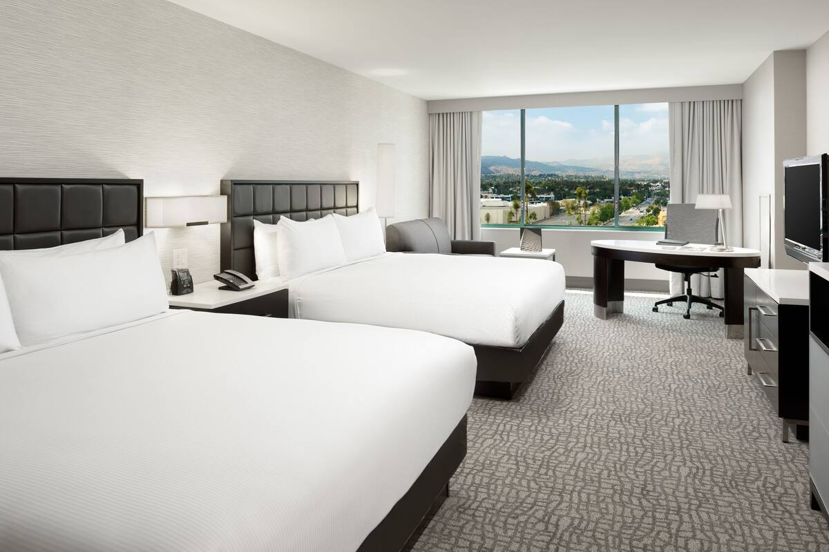 Habitación de hotel con dos camas, un escritorio, un televisor y un ventanal con vistas a la ciudad.