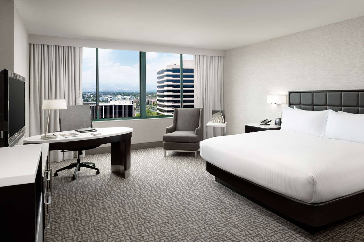 Habitación de hotel con una cama grande, un escritorio con silla, televisión y vista a un paisaje urbano a través de amplias ventanas.