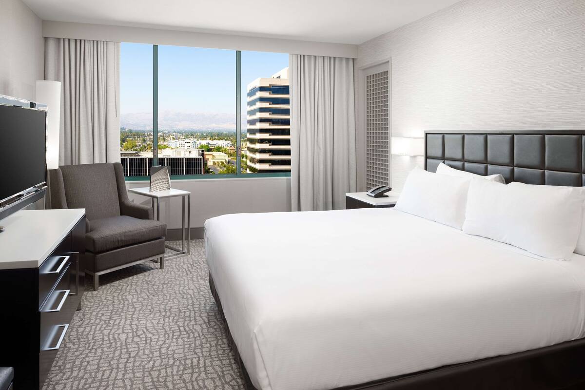 Una habitación de hotel con una cama grande, un escritorio, una silla, un televisor y vistas a un paisaje urbano a través de un ventanal amplio.
