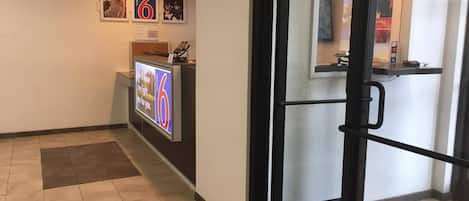 A hotel reception area with a digital screen displaying a message, a reception desk, and a glass door leading inside.