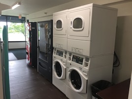 A laundry facility with stacked washers and dryers, a door leading outside, and a trash bin.